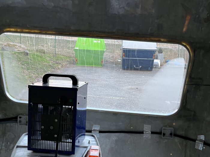 Värmefläkt inuti en vagn används för att torka ut fukt från ett fönster, synlig utsikt med en container och en sopcontainer utanför.
