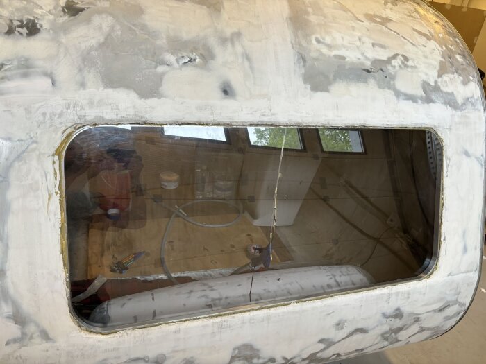 Bakfönster på en vagn med tydliga reparationer runt ramen, delvis slipat och spacklat. Inuti syns verktyg och slangar för pågående renovering.