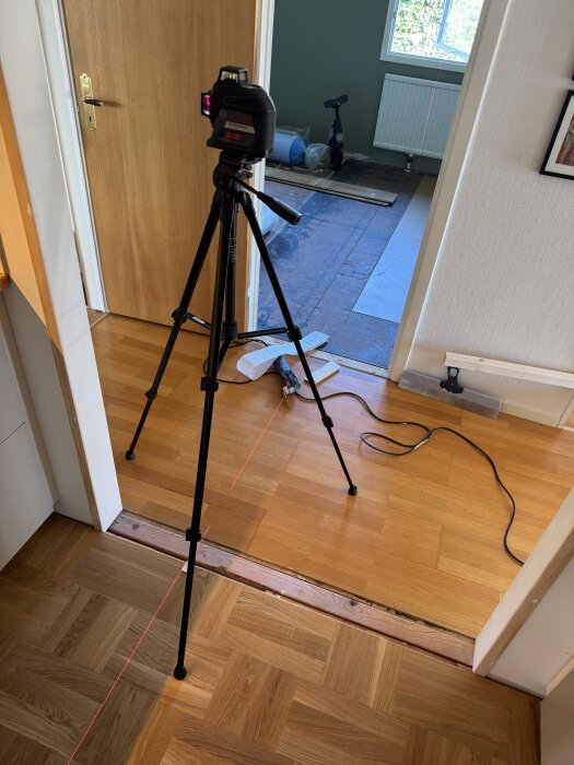 Laser på stativ används för att lägga golv i bösrummet, mönsterpassat med ekgolv, synlig fog mellan rum; arbetet sker under renovering.