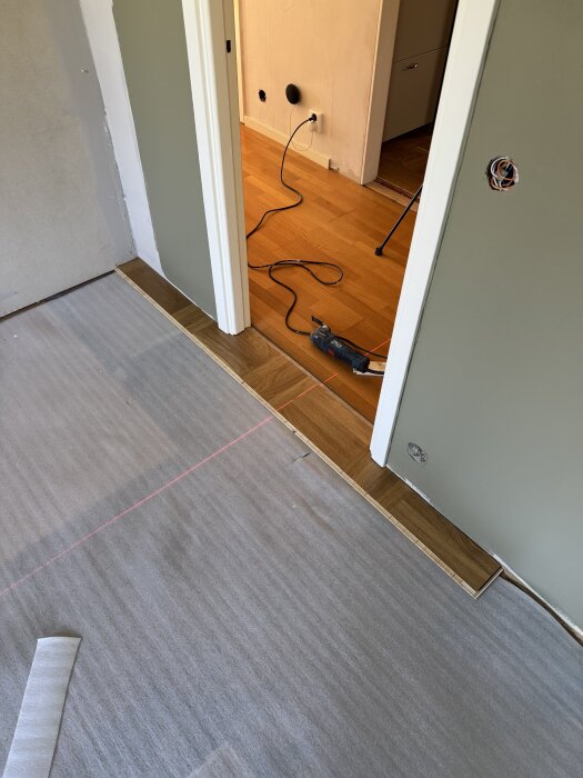 Laserlinje på golvet för att passa in ekmönsterbrädor mellan rum, halvlagda golvbrädor syns, verktyg på golvet och elkabel i hallen.