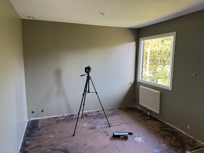 Tomt rum med gröna väggar, en laser på stativ, ett fönster och en radiator, förberedelser för golvläggning syns.