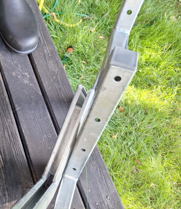 Metallvangstycke på en träaltan bredvid gräs, med flera hål för montering av trappsteg. Sulan av en stövel syns i bilden.