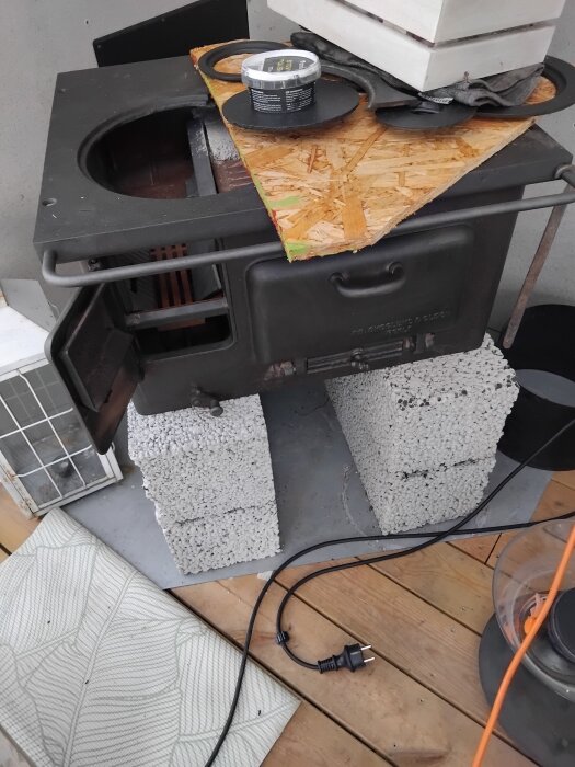 Gammal vedspis från Skoglund & Olsson, placerad på mursten, med öppen lucka och synligt fäste med glipa, samt elektrisk kabel framför.