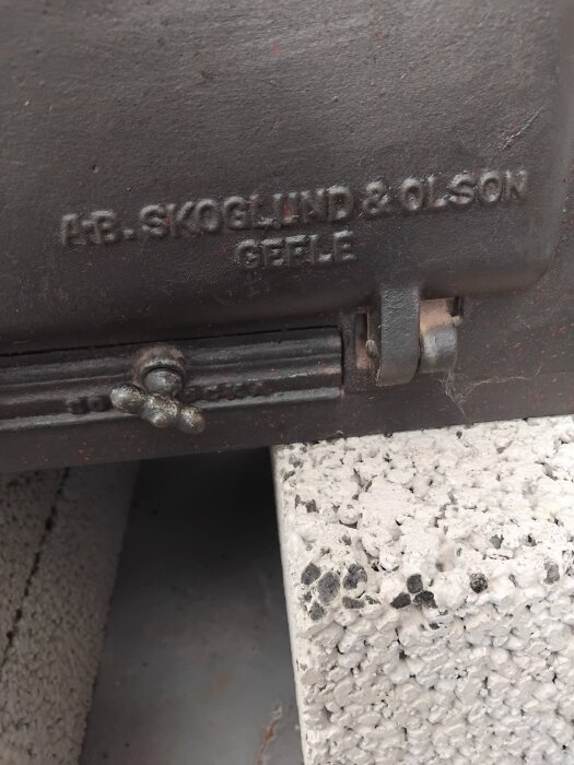 Gammal vedspis med texten "A.B. Skoglund & Olson, Gefle" på sidan, placerad på en mursten.