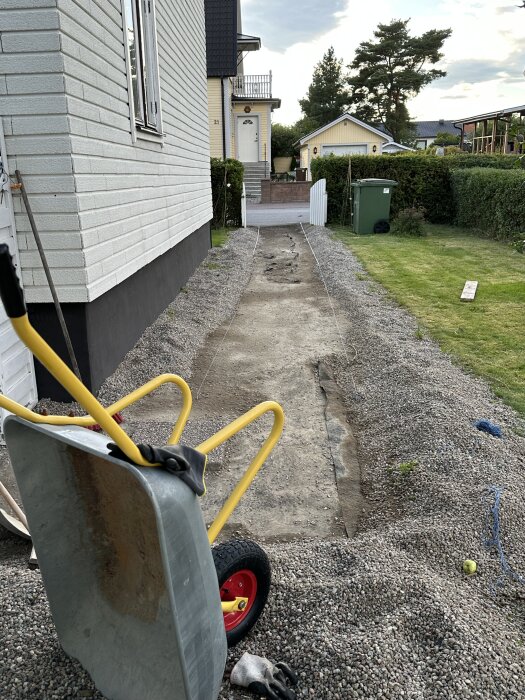 Grop för marksten med snören för breddmarkering, äldre markduk och gruskanter. Skottkärra och handskar syns, omgiven av hus och gräs.