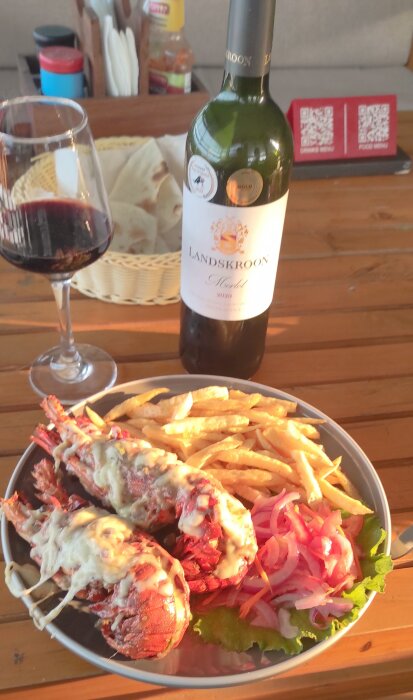 Languster med ost, pommes frites, och rödlök på tallrik, med Landskroon Merlot och rödvinsglas på bordet.