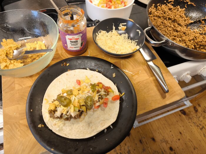Taco med fyllning av köttfärs, paprika, ost och jalapeños på tallrik bredvid tacochips, tacosalsa och riven ost i skålar.