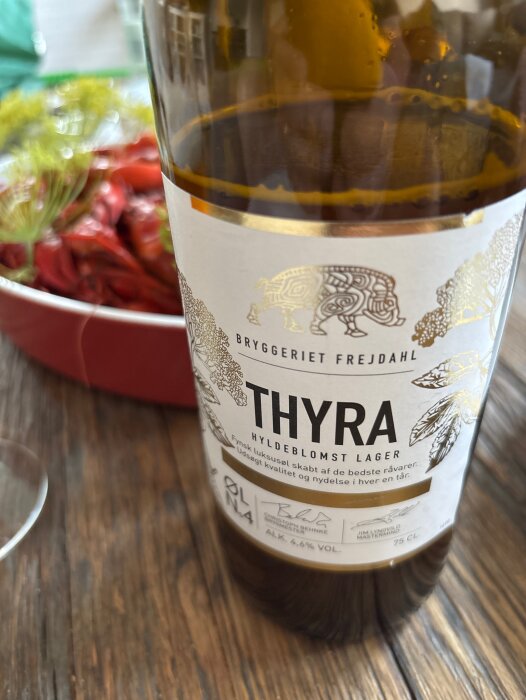 En flaska Thyra hyldeblomst lager från bryggeriet Frejdahl, står på ett bord med kräftor i bakgrunden.