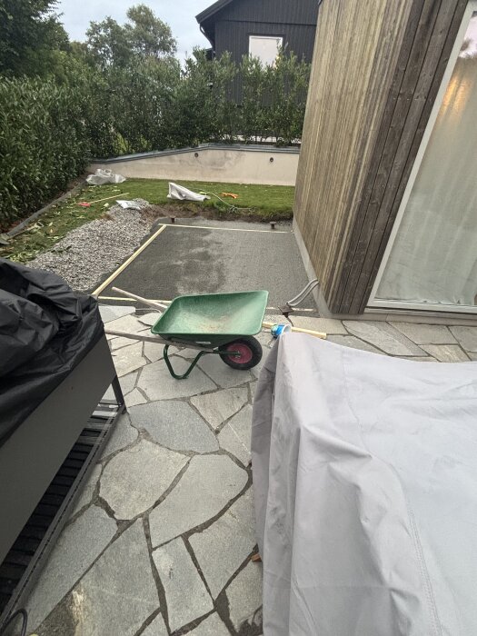 Grus- och betongyta framför hus med skottkärra, trädgårdstäppa och buskar. Underlag för kommande bygge av vardagsrumstillägg.