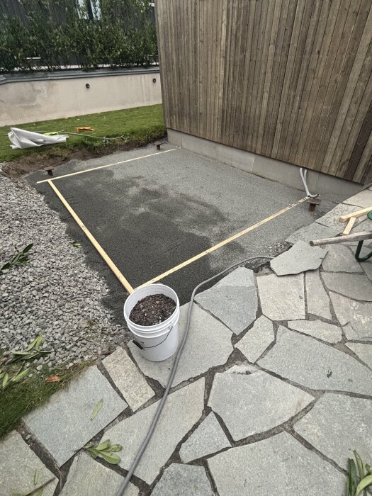 Byggarbetsplats med stockad och förberedd yta för platta, omgiven av träpanel, stenar och grus; vit hink i förgrunden.