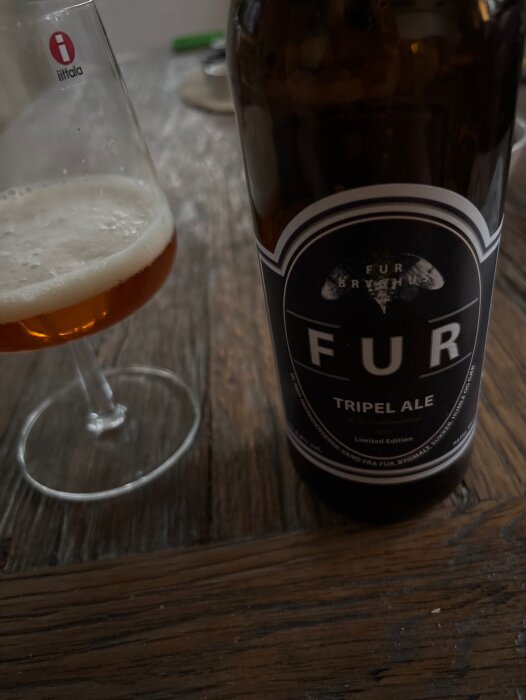 Flaska med Fur Tripel Ale bredvid ett glas med öl på träbord.