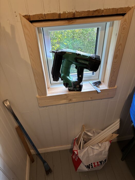 Fönster med ny träfoder, Hitachi spikpistol på fönsterbrädan, kofot mot väggen och en påse med träbitar på golvet.