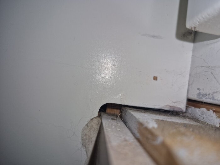 Springa under fönsterbräda mot gipsvägg, med synliga reglar och oregelbunden hålighet, omgiven av vitmålad träyta och viss smutsighet.