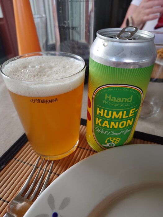 Ölglas bredvid en burk av Haand Bryggeriets Humlekanon West Coast IPA, och en gaffel på en bordstablett.