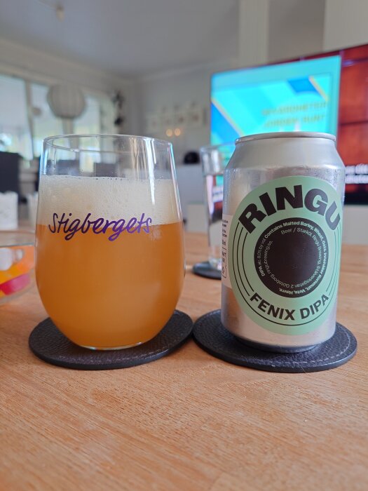 Glas med Stigbergets öl och burk med Ringu Fenix DIPA på ett bord, suddig bakgrund med TV.