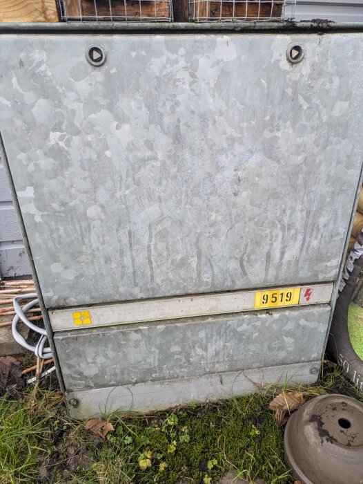 Grått elskåp nära fasaden av ett hus, med gräs nedanför och kablar synliga på sidan. Skåpet har en gul lapp med nummer "9519".