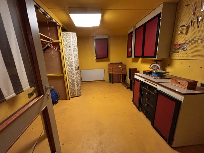 Verkstadsliknande källarrum med gul vägg och golv, rött och svart skåp, klädstång i dörröppning och olika verktyg på en perforerad väggtavla.