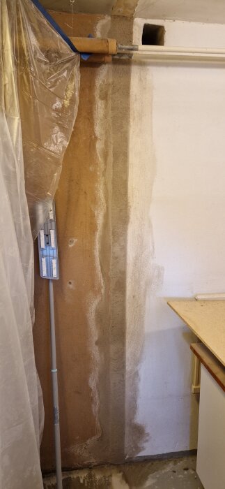 Vägg med olika putsytor i tvättstuga under renovering, plastskydd över rör och golv, väggpanel med synliga ojämnheter och strukturer.