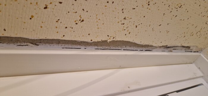 Vägg med sprucken puts och fläckig vävtapet ovanför en vit dörrlist i en tvättstuga under renovering.