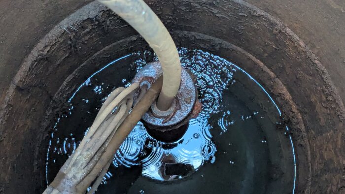 Närbild av ett gammalt rostfärgat brunnslock med kopplingar och vatten med bubblor, sett uppifrån i en brunn med rör och kablar.