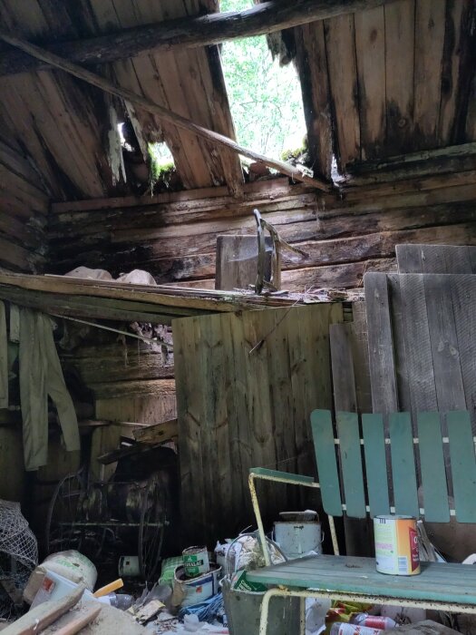 Förfallet uthus med trasigt tak, inredning synlig inklusive gammal stol, verktygshinkar och annan förvaring.