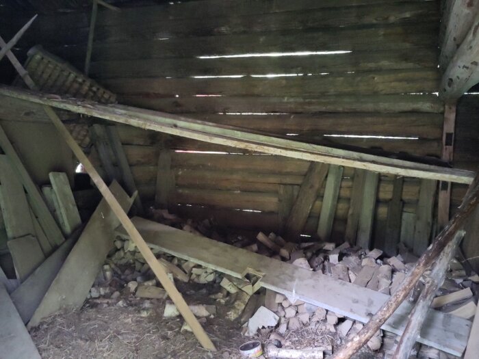 Inuti ett gammalt timrat uthus med rasade brädor, stockar och bråte, oskyddade väggar med ljusinsläpp genom springor i träet.