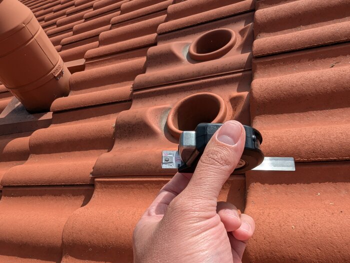 Hand håller ett måttband på ett tak med tegelpannor, jämte ett rör och två hål.
