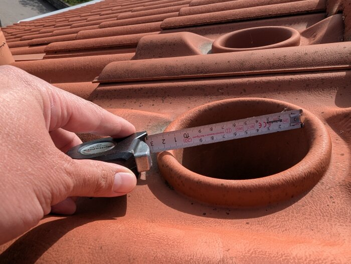 En tumstock mäter diametern på ett rött keramiskt takrör till 113 mm, mot en bakgrund av tegelpannor.