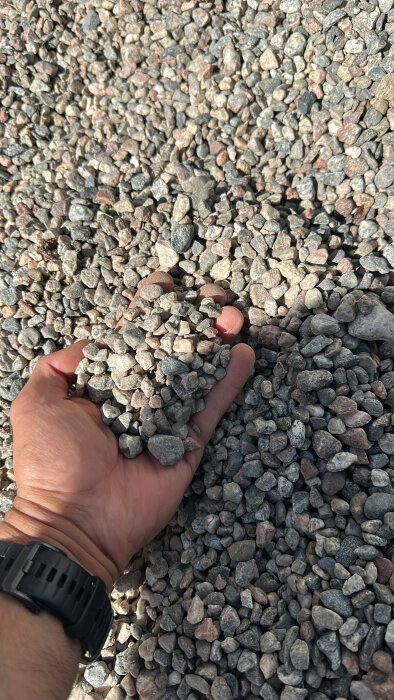 En hand håller i små grusstenar som ligger på en större hög, troligen använd som bärlager i byggprojekt.