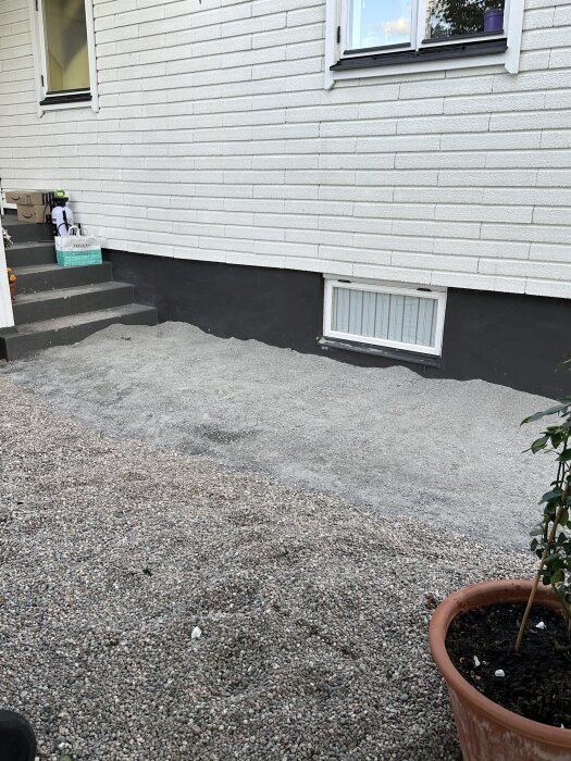 Grus ligger framför en vit putsad husvägg, med en krukväxt i förgrunden och en liten trappa till vänster.