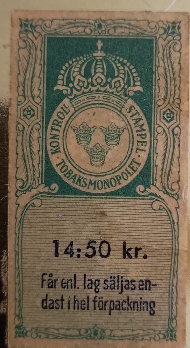 Etikett med texten "Kontrollstämpel Tobaksmonopolet" och priset 14:50 kr, ifrån en gammal cigarettask i plåt av märket John Silver.