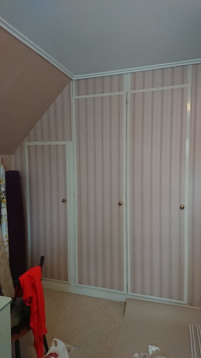 Garderobsvägg med tre dörrar och mönstrad tapet i ett rum, planeras för renovering och installation av pardörrar.