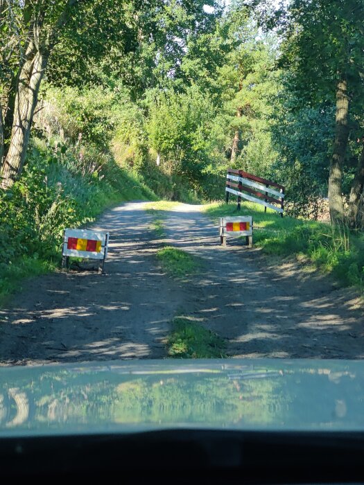 Smal grusväg med två väghinder i röd och gul färg. Vegetation omger vägen, och en vit grind syns i bakgrunden.