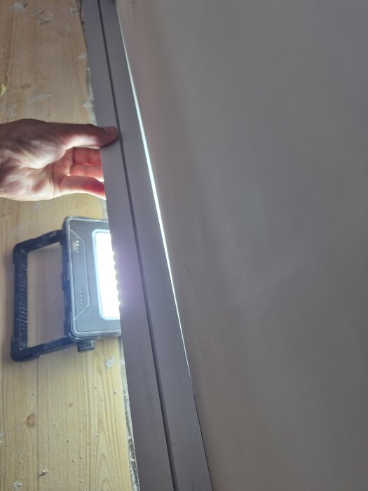 Hand som håller en tapetlinjal mot en vägg för att visa nivåskillnader, med en lampa i bakgrunden.