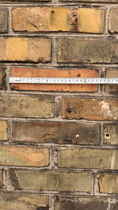 Gulbrunt tegel med måttband över, visar detaljer och slitage på fasaden.