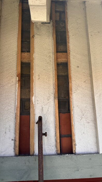 Vägg med träpanel borttagen visar tegel bakom, delvis dolt av vit puts och träreglar, med en rostig metallstång i förgrunden.