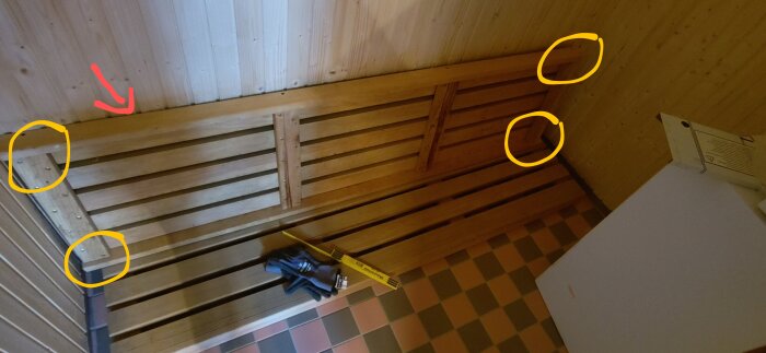 Bastulavar av trä i ett hörn med markerade områden för fästen, en röd pil pekar på en bräda och det finns mätverktyg och handskar på laven.