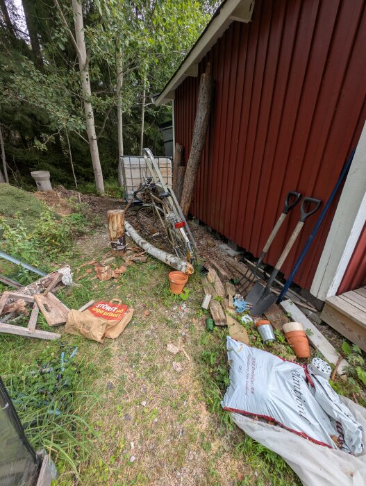 Röd verktygsbod med löv och skräp runtom; cykel, ved, verktyg och träd i bakgrunden. Grus och jord på marken.