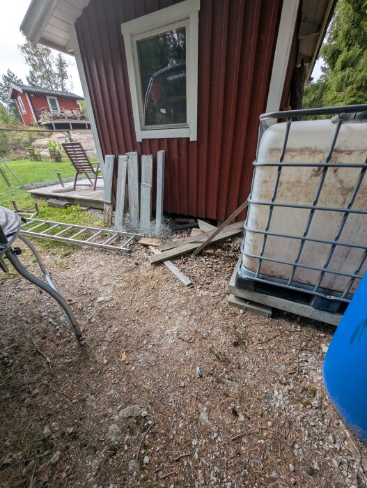 Förråd med röd fasad, diverse brädor och material på marken och en vattentank bredvid, omgiven av naturlig terräng och en ladderspärr vid sidan.