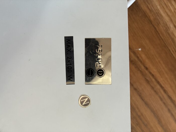 Skyltar med texten 'MAX 2x100 W', '75202', symboler och bokstaven 'N' på en vit yta, relaterat till taklampa och elkoppling.