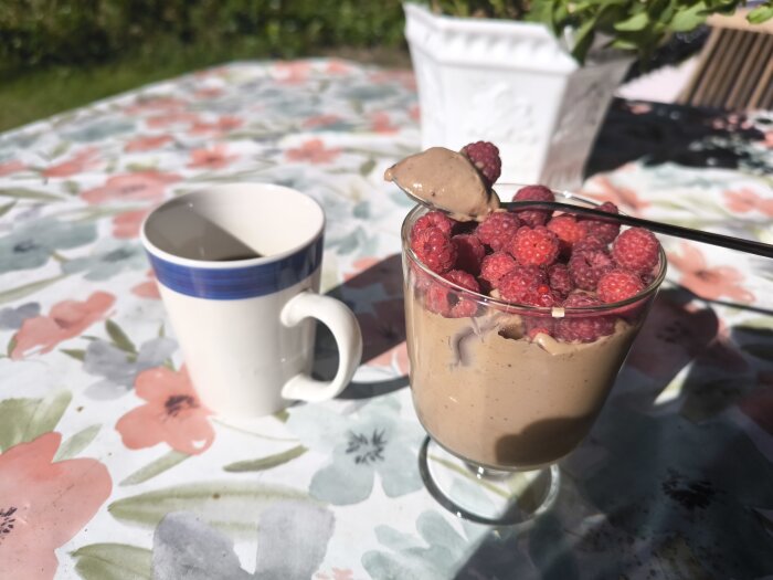 Toblerone-mousse toppad med färska hallon i ett glas bredvid en kaffekopp på ett blommigt bord.