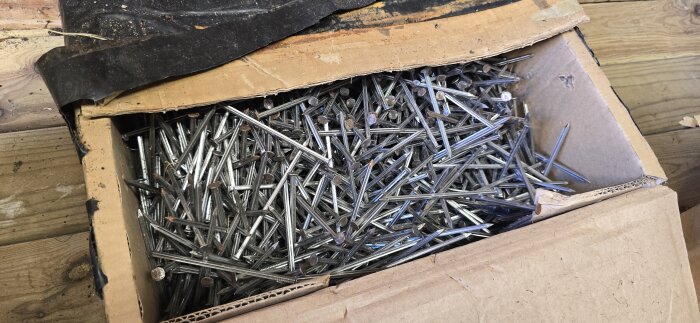 En öppen kartong fylld med många metallspikar, stående på en träyta.