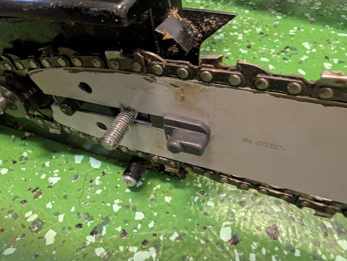 Motorsågens kedja och svärd med justeringsbult på ett grönt golv.