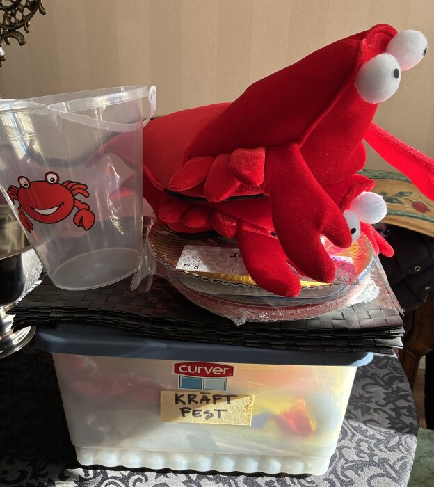 Kräftskiva-förberedelser med plastkrabba, dekorativa kräftor, en plastmugg med krabbmotiv och en låda märkt "KRAFTFEST" på ett bord.