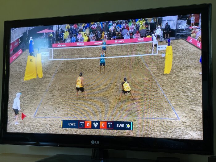 Beachvolleybollmatch visas på TV med två spelare i gula tröjor på ena sidan och en spelare i turkos tröja på andra sidan och publik i bakgrunden.