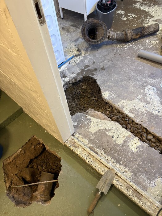 Gjutjärnsrör borttaget från betonggolv, grävt dike syns. Spade och verktyg ligger vid sidan. Reparation av avloppssystem i källare.