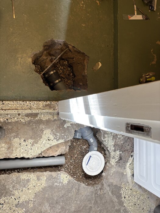 Bild av en nyinstallerad avloppsbrunn med rör och kopplingar i en gjuten golvyta, omgiven av jord och grus.