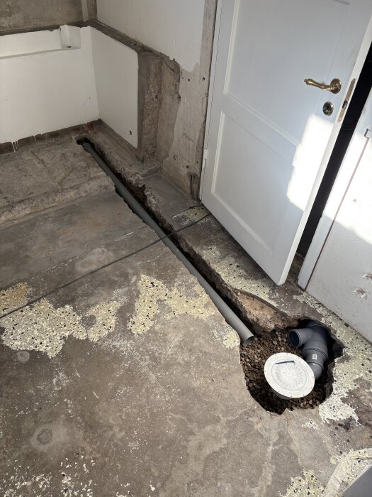 Renovering med ny avloppinstallation: öppnat golv med synliga rör och brunn, dörr i bakgrunden, del av projektet att ersätta gammalt gjutjärnsrör.