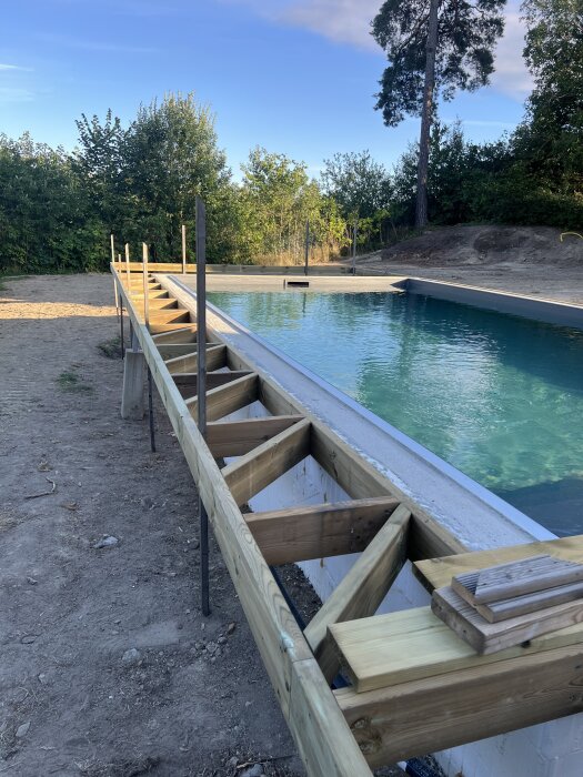 Byggarbetsplats med en oavslutad ramen runt en pool i en trädgård, skog i bakgrunden och klarblå himmel ovanför.