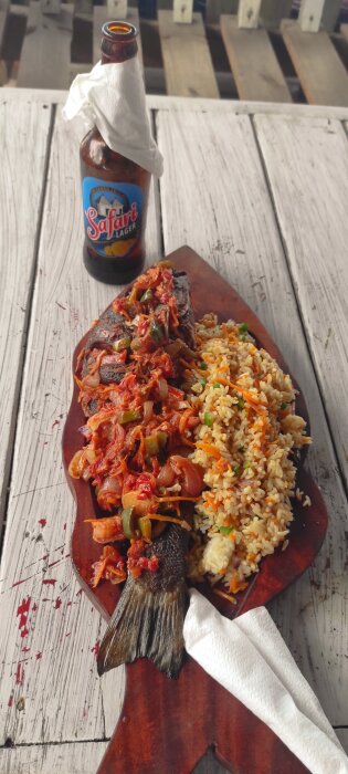 Tillagad fisk med sås och ris på en träbricka, serverad med en flaska Safari Lager på ett träbord.